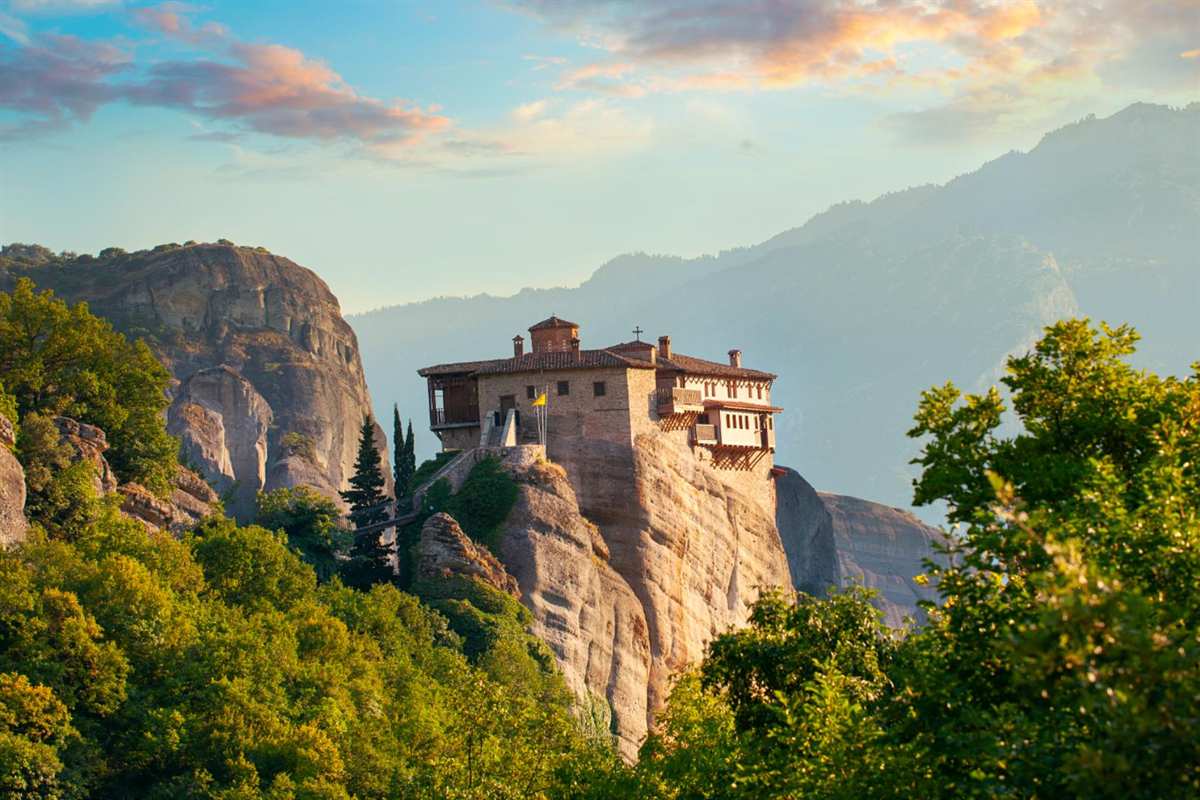 Greece tours: a Meteora monastery with terracotta roofs and wooden balconies perched on a tall sandstone rock pillar, surrounded by green trees, with hazy mountains and a pale blue sky with pink clouds behind it