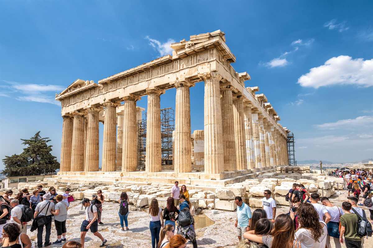 Greece tours: the Parthenon on the Acropolis in Athens, its white marble columns and roof lit by sunshine under a clear blue sky with scattered white clouds, and visitors standing on the stone ground in front